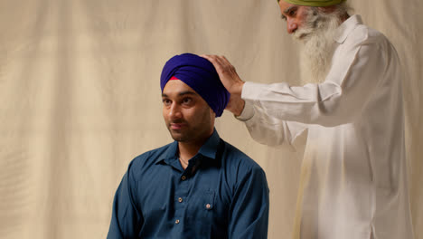 Studioaufnahme-Von-Zwei-Sikh-Männern,-Die-Beim-Aufsetzen-Eines-Turbans-Vor-Einem-Einfachen-Hintergrund-Eine-Salai-Nadel-Verwenden